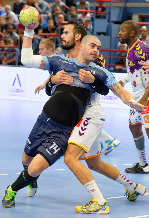 Eine Gruppe von Männern, die Handball in einem Stadion spielt, wobei ein Spieler den Ball hält.