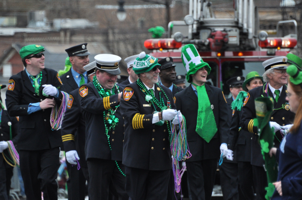 Eine Gruppe uniformierter Männer marschiert in einem St. Patrick's Day-Umzug, hält Perlen und hat ein Feuerwehrauto und Gebäude im Hintergrund.