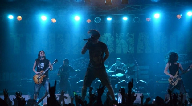 Eine Gruppe von Musikern auf einer hell erleuchteten Bühne mit sichtbarem Publikum, mit Gitarristen, einem Schlagzeuger und einem Sänger mit Mikrofon.