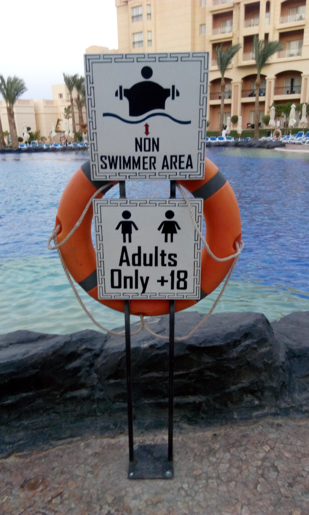 Ein Schwimmbecken mit einem "Non Swimmer Area Adults Only"-Schild im Vordergrund, umgeben von Bäumen und Gebäuden unter einem klaren blauen Himmel, mit einem Rettungsring in der Mitte des Pools.