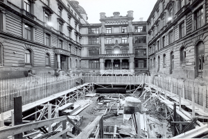 Schwarzes und weißes Foto einer Baustelle in einer Stadt mit Gebäuden im Hintergrund, verstreute hölzerne Gegenstände und Metallstangen im Vordergrund und Text unten rechts.