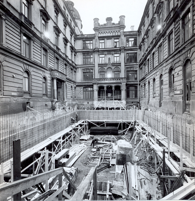 Schwarzes und weißes Foto einer Baustelle in einer Stadt mit Gebäuden im Hintergrund, verstreute hölzerne Gegenstände und Metallstangen im Vordergrund und Text unten rechts.