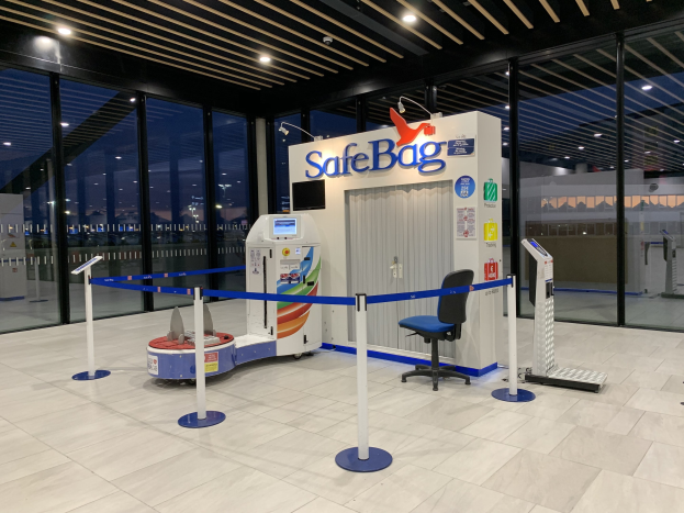 Sicherheitskontrolle für Gepäck am Flughafen mit einem Stuhl, Absperrböcken, Maschinen, einem Bildschirm, einer bedruckten Tafel, Glaswänden, Deckenleuchten und sichtbarem Boden.