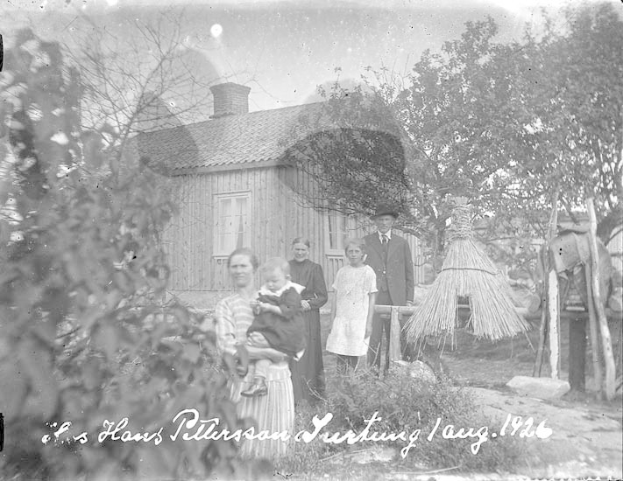 Ein altes Schwarz-Weiß-Foto einer Familie, die vor einem Haus steht, mit einer Frau, die ein Baby hält, anderen Familienmitgliedern drumherum, Bäumen, einer Hütte im Hintergrund und Text unten.