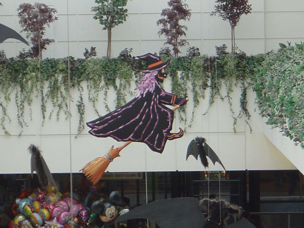 Halloween-Umzug mit Hexe auf Besen vor einem Gebäude, mit Spielzeug und Luftballons im Vordergrund, einer bepflanzten Wand im Hintergrund und einem Geländer auf der rechten Seite.