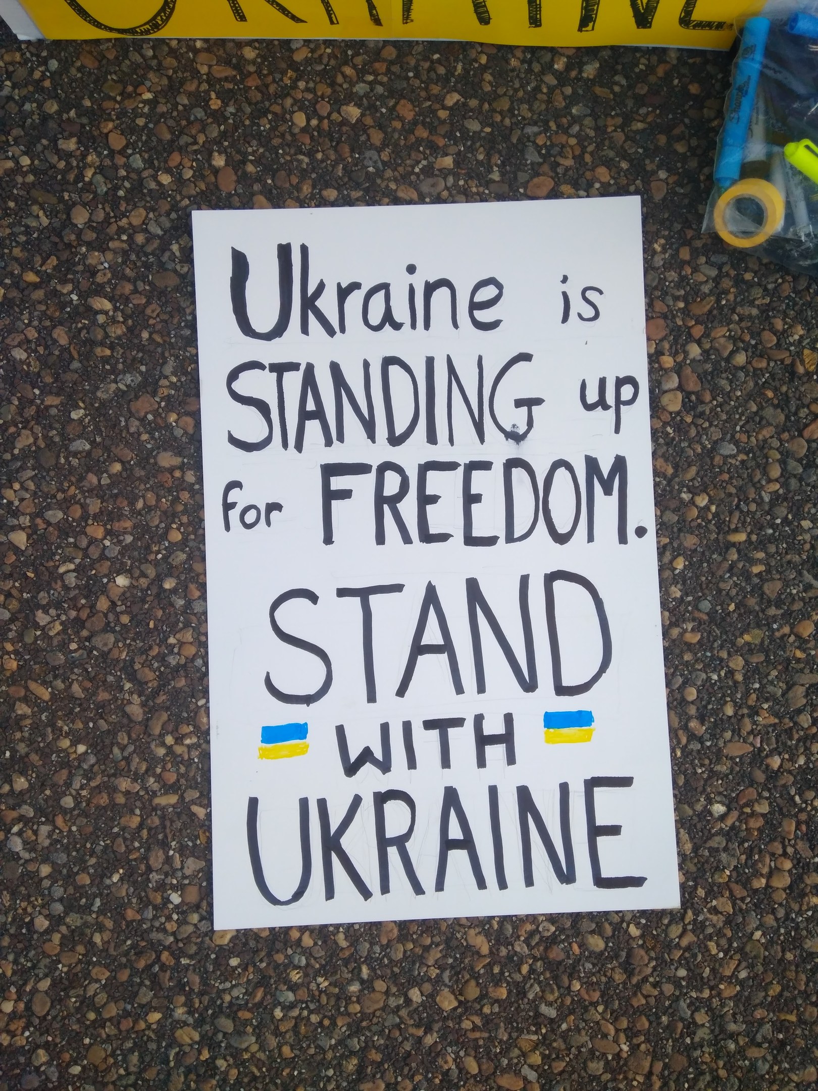 Ein weißes Blatt Papier mit dem Text "Ukraine steht für Freiheit ein, steht mit Ukraine" liegt vor einer gelben Fahne auf dem Boden. Rechts davon befindet sich eine Plastikhülle mit verschiedenen Gegenständen.