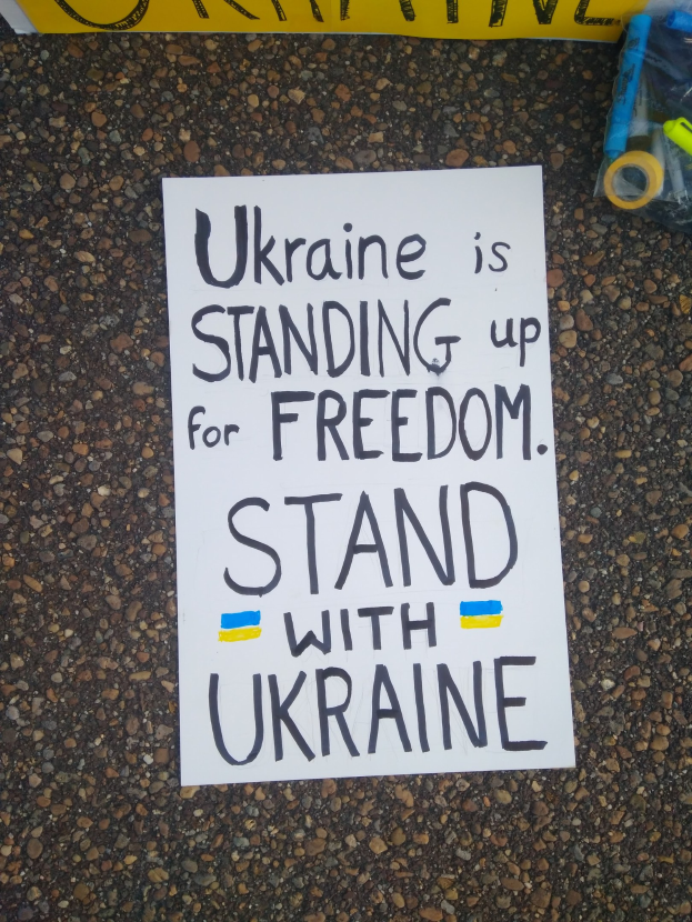 Ein weißes Blatt Papier mit dem Text "Ukraine steht für Freiheit ein, steht mit Ukraine" liegt vor einer gelben Fahne auf dem Boden. Rechts davon befindet sich eine Plastikhülle mit verschiedenen Gegenständen.