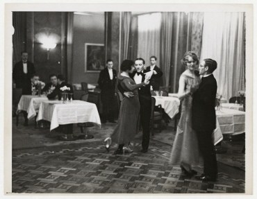 Schwarzes und weißes Foto eines Paares, das in einem Ballsaal tanzt, mit Zuschauern an Tischen und Vorhängen im Hintergrund.