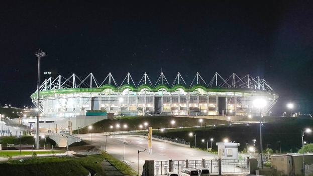 Ein großes Stadion bei Nacht beleuchtet mit parkenden Autos davor, umgeben von Straßenmasten, Straßenlaternen, Strommasten, Stromkabeln, Fahrzeugen auf der Straße, Gittern, Bäumen und einem sternenklaren Himmel.