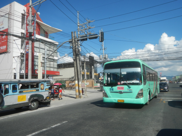 Ein grüner Bus fährt auf einer von Hochhäusern gesäumten Stadtstraße, während Passanten mit Regenschirmen auf dem Gehweg darunter gehen und Strommasten mit Drähten darüber verlaufen.