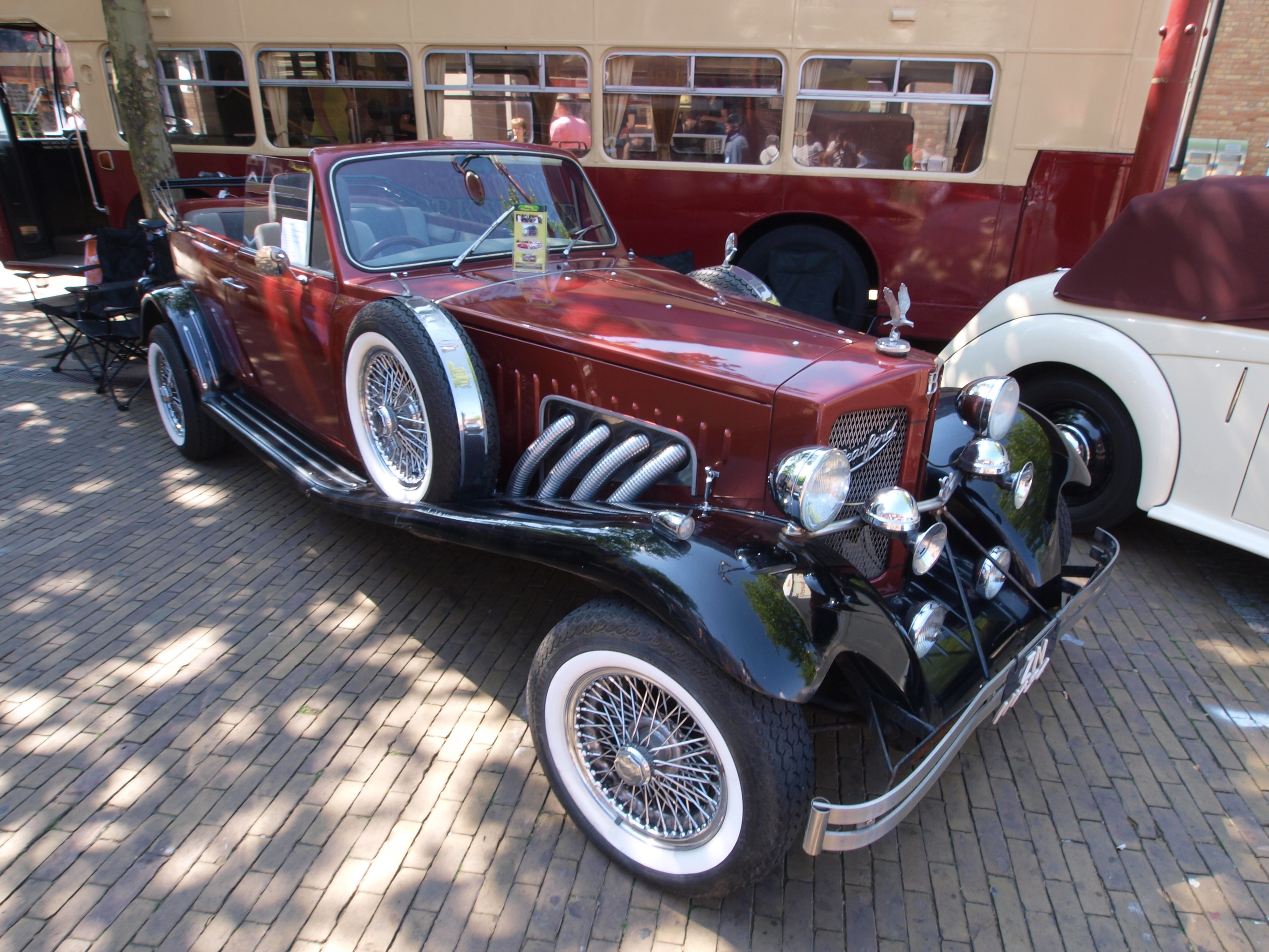 Ein Oldtimer neben einem Doppeldeckerbus auf einem Kopfsteinpflasterweg, mit Stühlen und einem Baum im Vordergrund, einem Gebäude im Hintergrund und Menschen im Bus.