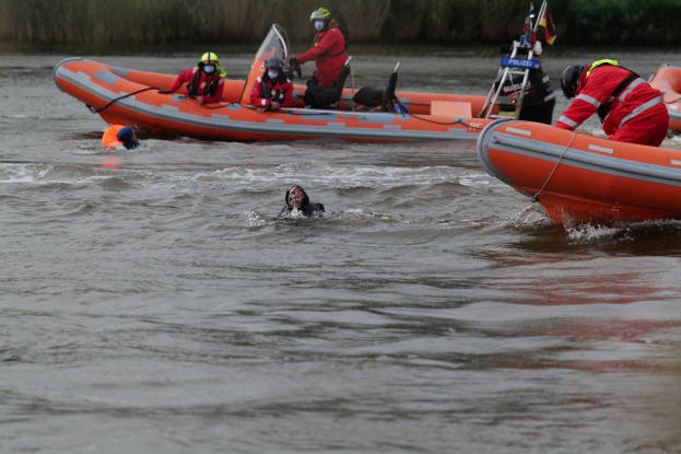 Gruppe von Menschen in einem aufblasbaren Boot auf einem Fluss, mit zwei Personen im Wasser im Vordergrund, die Lebenswesten und Helme tragen, und einigen Pflanzen im Hintergrund.