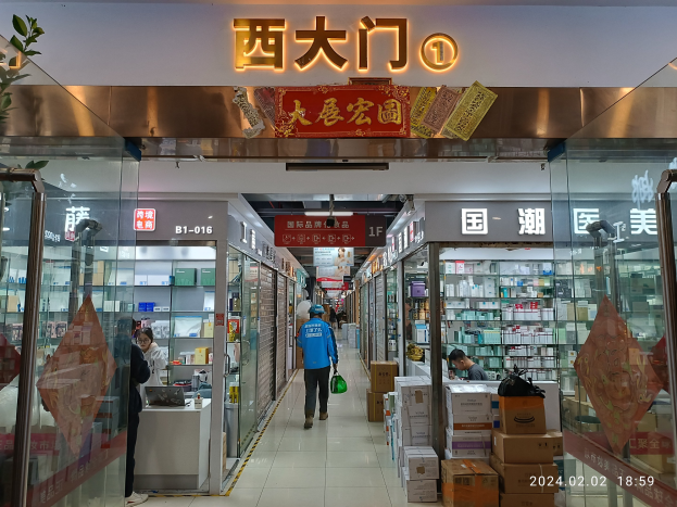 Innere eines chinesischen Apotheken mit Glaswänden, einer Pflanze auf der linken Seite, Regalen voller Gegenstände, verstreuten Kartons auf dem Boden und einem Namensschild oben.