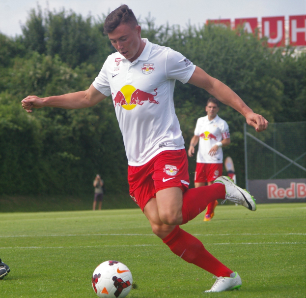 Ein Profifußballer schießt einen Ball auf einem grünen Feld mit Bäumen und einem klaren blauen Himmel im Hintergrund, in der Nähe einer Tafel mit der Aufschrift "RB Leipzig."