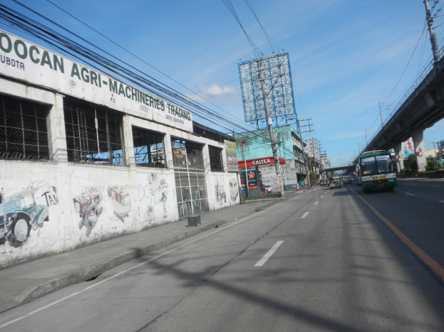 Stadtstra├če mit fahrenden Fahrzeugen, eine Brücke auf der rechten Seite, Geb├Ąude, Strommasten mit Kabeln, ein bew├Âlktes Himmel und ein graffiti-beschmiertes Geb├Ąude an der Seite.