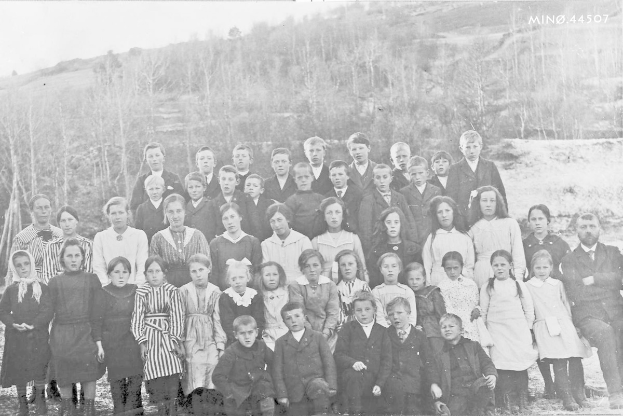 Schwarzes und weißes Foto einer Gruppe von Kindern, die vor einem Hintergrund aus Bäumen und einem Hügel posieren, mit der Beschriftung "1930-1940 Schulkinder" unten.