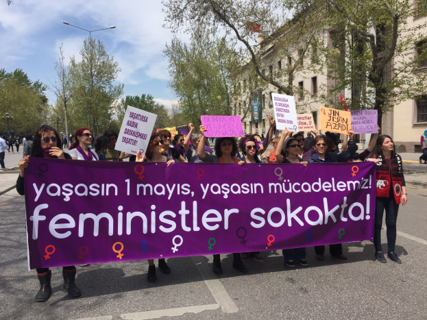 Eine Gruppe von Menschen mit einem violetten Banner mit der Aufschrift 'feministische sokokta' geht eine von Bäumen gesäumte Straße mit Laternenpfählen und einem Gebäude im Hintergrund unter einem bewölkten Himmel entlang.