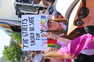 Eine Gruppe von Menschen steht vor einem Lastwagen und hält ein Schild hoch, auf dem "Die Straßen sollten sicher sein" steht. Einige der Menschen tragen Mützen und Schutzbrillen, andere halten Schilder. Im Hintergrund sind Bäume, Gebäude und ein klarer blauer Himmel zu sehen.