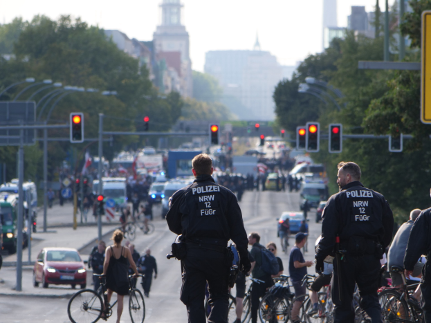 Eine Gruppe von Polizisten auf Fahrrädern, die eine von Bäumen gesäumte Straße mit Gebäuden und einem klaren blauen Himmel im Hintergrund entlangfahren.