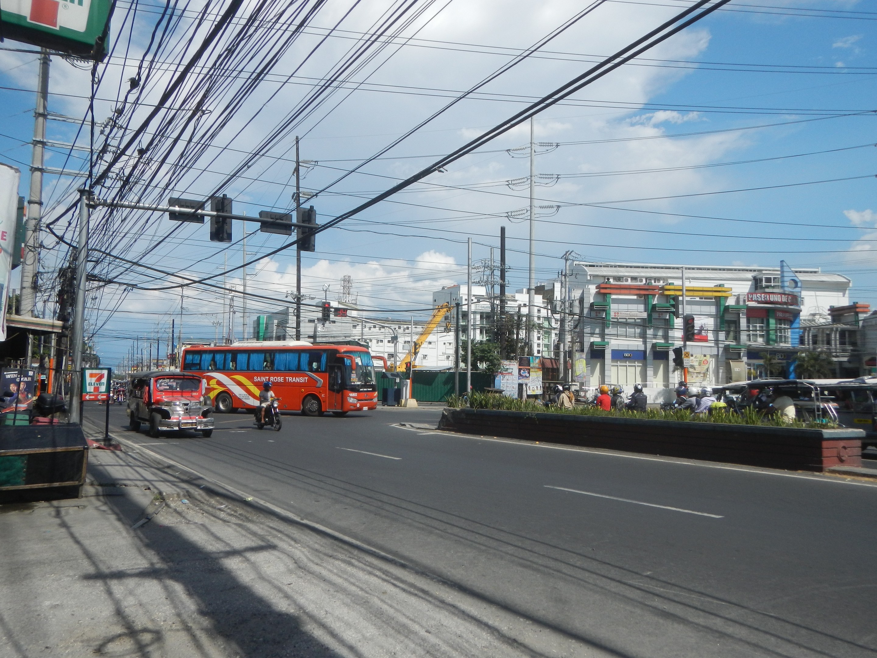 Eine belebte Stadtstraße mit Fahrzeugen, Strommasten, Verkehrsampeln, Schildern, Bäumen, Gebäuden und einem bewölkten Himmel.