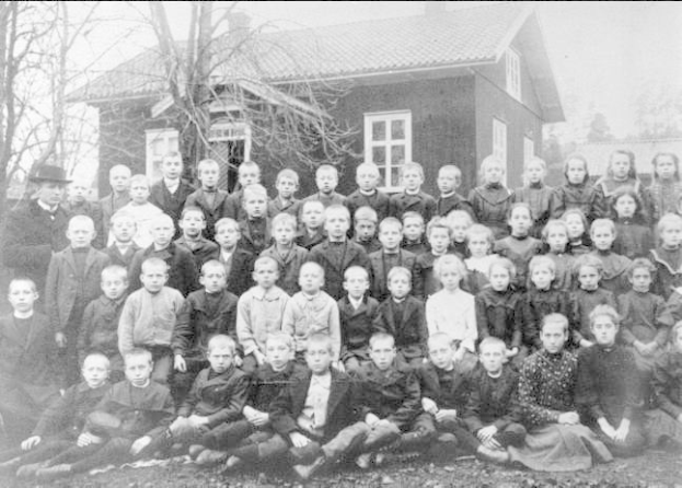 Schwarz-weißes Foto einer Gruppe von Kindern, die vor einem Haus mit Fenstern posieren, umgeben von Bäumen und einem klaren Himmel, mit dem Text "1930er Schulkinder" unten.