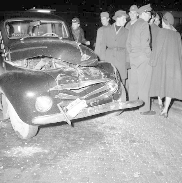 Schwarzes und weißes Bild eines schwer beschädigten Autos am Straßenrand mit einer Gruppe von Menschen, die es inspizieren.
