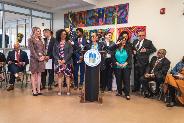 Eine Gruppe von Menschen steht vor einem Rednerpult mit Mikrofon und einer Tafel mit der Aufschrift "Sancho Stadtrat tritt dem Stadtrat bei", im Hintergrund sitzen und stehen andere, Fahnen, eine Tafel, ein Feuerlöscher und Deckenlampen an der Wand.