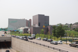 Stadtstraße mit fahrenden Autos, eine Brücke mit Geländern, Laternen, Verkehrsampeln an Masten, Bäume, Gebäude mit Fenstern und ein sichtbarer Himmel.