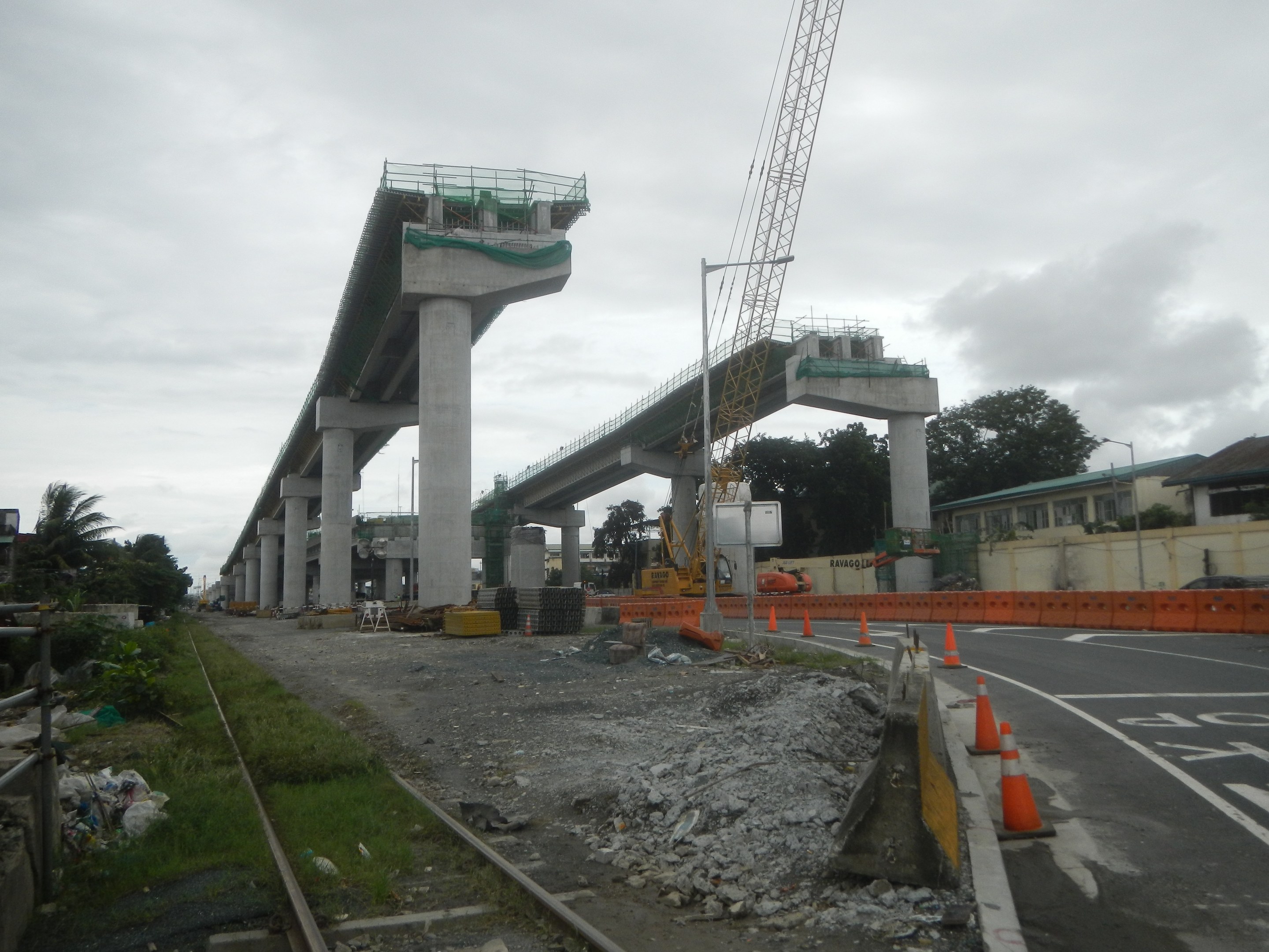 Baustelle mit einer Brücke im Hintergrund, Straße mit Absperrkegeln markiert, Bahnschiene links, verstreute Steine und Gras, Bäume und Gebäude säumen die Straße und ein bewölkter Himmel.