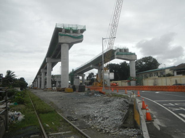 Baustelle mit einer Brücke im Hintergrund, Straße mit Absperrkegeln markiert, Bahnschiene links, verstreute Steine und Gras, Bäume und Gebäude säumen die Straße und ein bewölkter Himmel.