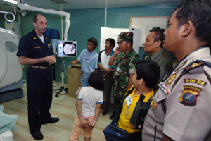 Eine Gruppe von Menschen steht in einem Krankenhauszimmer, ein Mann hält ein Mikrofon in der Mitte, links ein Bett mit einer Decke darauf und verschiedene medizinische Geräte, ein Röntgengerät, eine Tafel mit Text, eine Tür, eine Wand und eine Decke mit Lampen im Hintergrund.