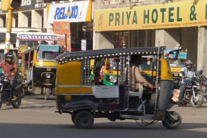 Eine belebte Straße mit verschiedenen Fahrzeugen, einschließlich eines Auto-Rikschas, und Menschen mit Helmen, mit einer Ampel im Vordergrund und Gebäuden mit Namensschildern und Rollläden im Hintergrund.