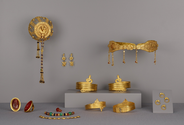 Goldschmuck-Präsentation mit Armbändern, Ringen und weiteren Stücken auf einer Oberfläche, mit zusätzlichen Teilen an der Wand im Hintergrund.