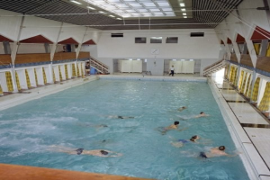 Großes Hallenbad mit Schwimmern, Säulen auf beiden Seiten, einer Uhr an der Wand im Hintergrund und Deckenbeleuchtung.