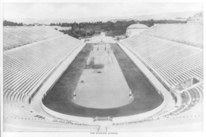 Schwarzes und weißes Foto des Olympischen Stadions in Athen, Griechenland, mit Sitzreihen, umgebenden Bäumen, fernen Gebäuden und Bergen, mit Text unten.