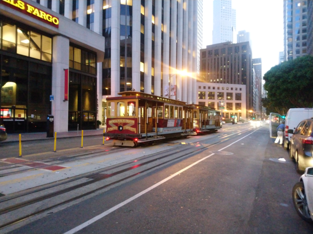 Seilbahn auf einer Stadtstraße in San Francisco mit Fahrzeugen, Fußgänger, parkendem Van, Gebäuden, Bäumen und Himmel im Hintergrund.