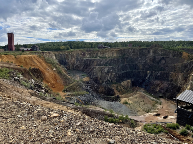 Großer Tagebau umgeben von Bäumen, Gebäuden und einem Schuppen, mit bewölktem Himmel und Boden bedeckt mit Steinen und Pflanzen.