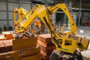 Großes Industriegebäude mit gestapelten Kisten, Kränen, Zaun, Stützpfählen und einer fernen Wand in einer Fertigungseinrichtung.