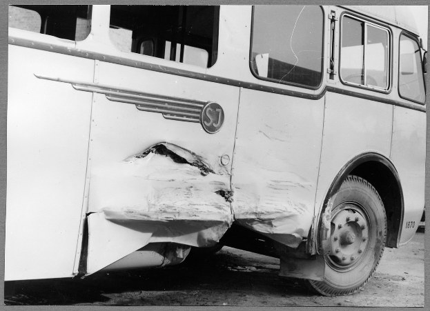 Schwarz-weißes Foto eines beschädigten Busses mit eingedellter Front, zersplitterten Fenstern und geöffneter Fahrertür am Straßenrand.