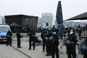 Eine Gruppe von Polizeibeamten steht vor einer Menge von Demonstranten in Berlin, Deutschland, mit Barrikaden, Zelten und Fahnen im Vordergrund und Gebäuden unter einem klaren Himmel im Hintergrund.