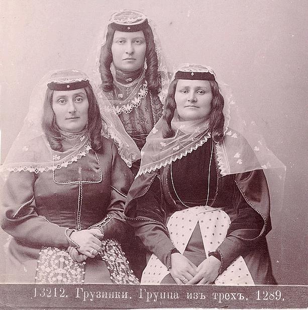 Drei Frauen in traditionellen russischen Hochzeitskleidern mit farbenfroher Stickerei und Perlen stehen vor einer einfarbigen weißen Wand in einem Schwarz-Weiß-Foto.
