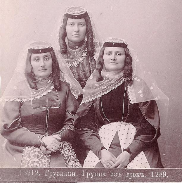 Drei Frauen in traditionellen russischen Hochzeitskleidern mit farbenfroher Stickerei und Perlen stehen vor einer einfarbigen weißen Wand in einem Schwarz-Weiß-Foto.