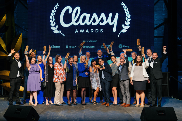 Gruppe von Menschen auf einer Bühne, die Auszeichnungen halten, mit Rednern darunter und einem Bildschirm im Hintergrund, der 'Classy Awards 2019' anzeigt.
