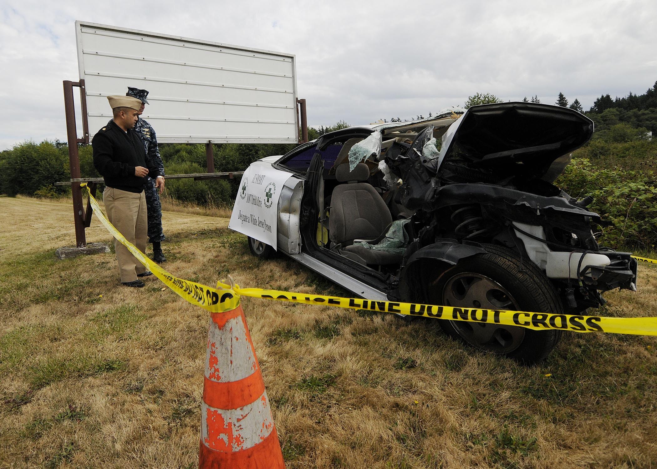 Ein Polizeibeamter in Uniform steht neben einem schwer beschädigten Auto am Straßenrand, mit eingedellter Motorhaube und zersplitterten Fenstern, neben einem Verkehrskegel und einem bandagierten Pfahl, vor einem Hintergrund aus Bäumen und einem bewölkten Himmel.