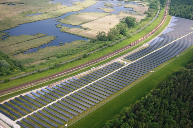Luftaufnahme eines Solarparks mit Panelen in einem Feld, umgeben von Bäumen, Gras und Wasser, mit einem Zug auf einer nahen Bahnstrecke.