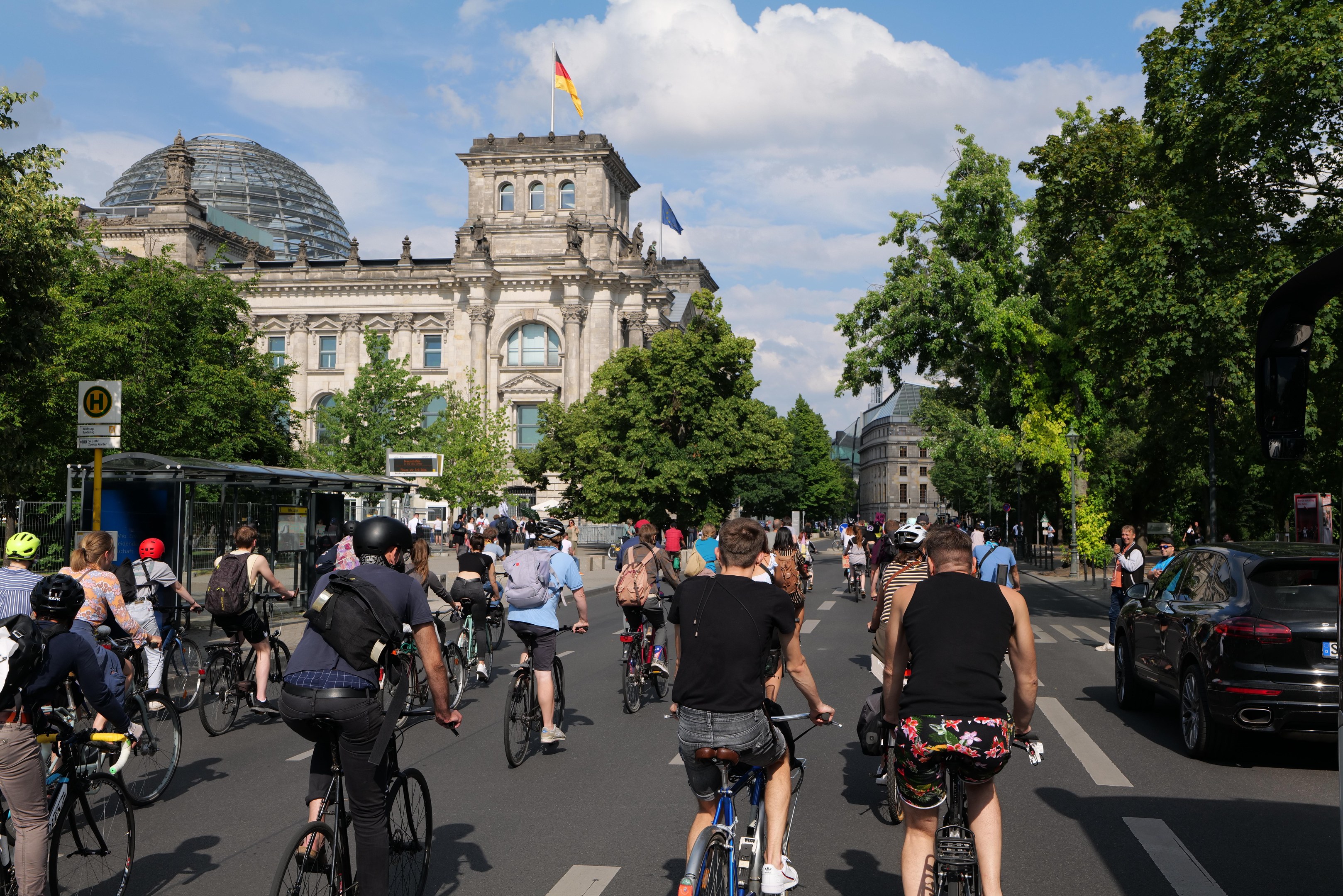 Eine Gruppe von Menschen, die auf Fahrrädern eine von Bäumen gesäumte Straße in Berlin, Deutschland, entlangfahren, mit einer Bushaltestelle auf der rechten Seite und einer Flagge, die auf einem Gebäude weht, unter einem bewölkten Himmel.