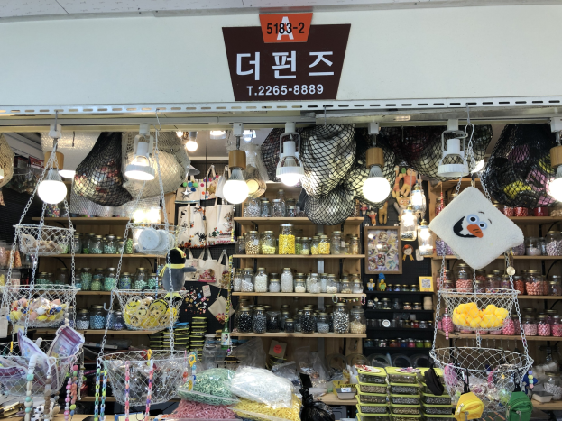 Ein unordentlicher Laden in Seoul, der verschiedene Gegenstände wie Körbe, Lichter, Flaschen und Spielzeuge auf Regalen ausstellt, mit einer Tafel öber der Tür, die ihn als bestbewerteten koreanischen Shop ausweist.