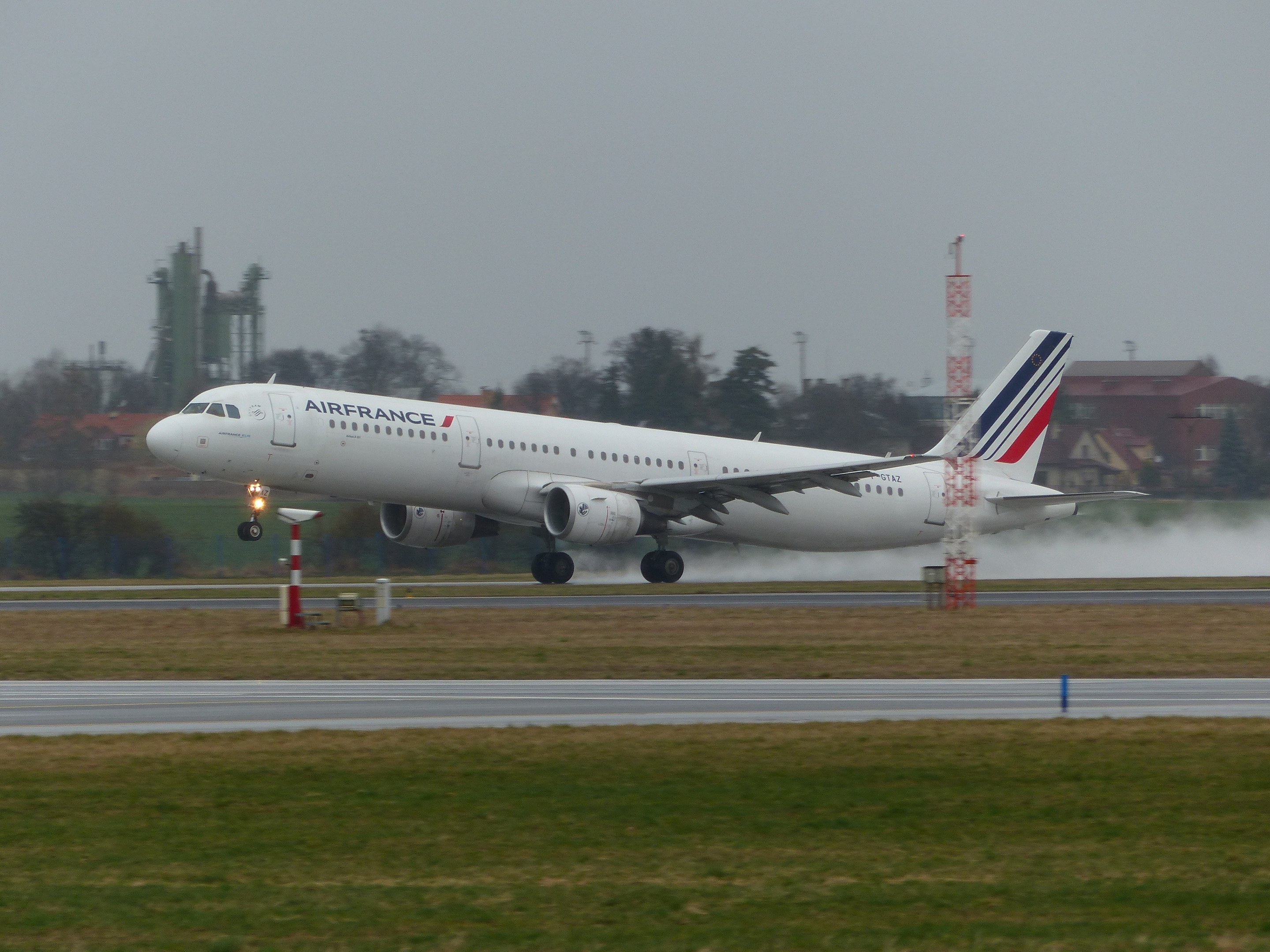 Air France Airbus A320-200 beim Abheben vom Pariser Charles-de-Gaulle-Flughafen, umgeben von grünem Gras und Bäumen, mit Gebäuden und Türmen im Hintergrund und einem klaren blauen Himmel.