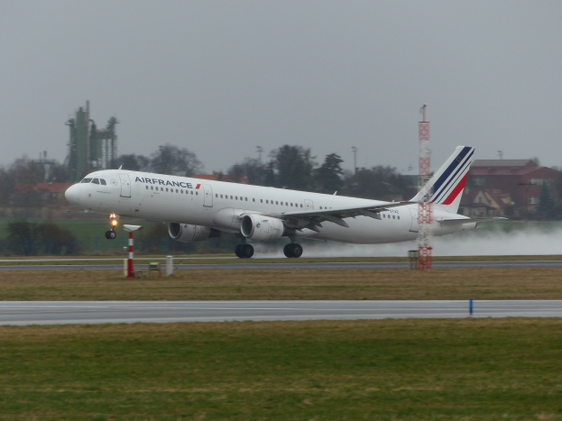 Air France Airbus A320-200 beim Abheben vom Pariser Charles-de-Gaulle-Flughafen, umgeben von grünem Gras und Bäumen, mit Gebäuden und Türmen im Hintergrund und einem klaren blauen Himmel.