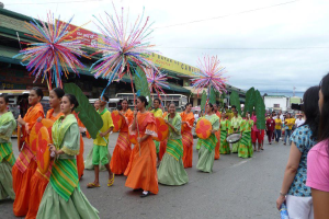 Eine Gruppe von Menschen in bunten Kostümen marschiert auf einer Straße während eines Karnevalsumzugs, wobei eine Frau rechts eine Flasche hält, und Fahrzeuge, Gebäude, Bäume, Pfosten, Drähte und der Himmel im Hintergrund zu sehen sind.